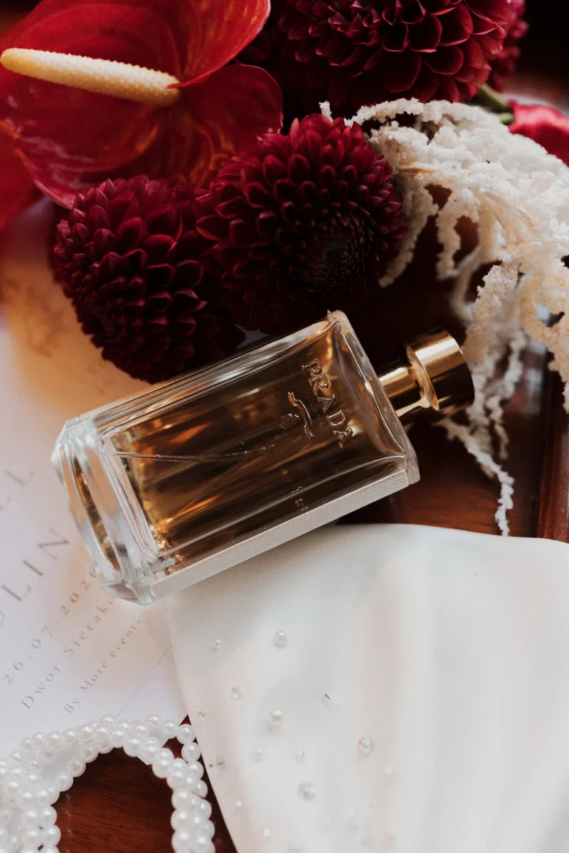 Perfumy i biżuteria panny młodej obok bordowych dalii i czerwonego anturium