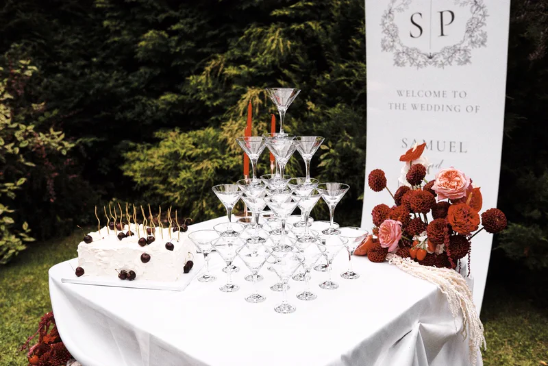 Wieża kieliszków i tort obok tablicy powitalnej, przy nich kompozycja z bordowych dalii